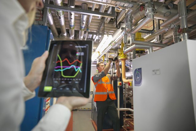 Office workers using digital tablet and camera to check efficiency of office heating in boiler room Unternehmen investieren verstärkt in den Klimaschutz und auch Verbraucher können von Energiesparpotenzialen profitieren.