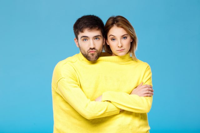 Couple in yellow sweater posing with crossed arms over blue background.