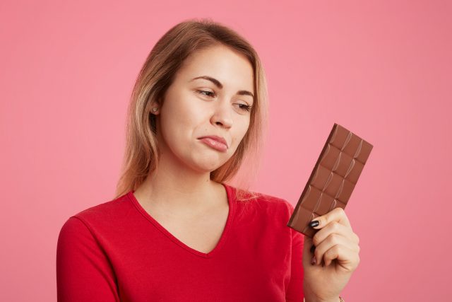 Attractive young woman looks with discontent expression at sweet bar of chocolate, keeps to diet, can`t eat it to be slim and sporty, curves lower lip, isolated over pink background. Eating sweets Schokolade