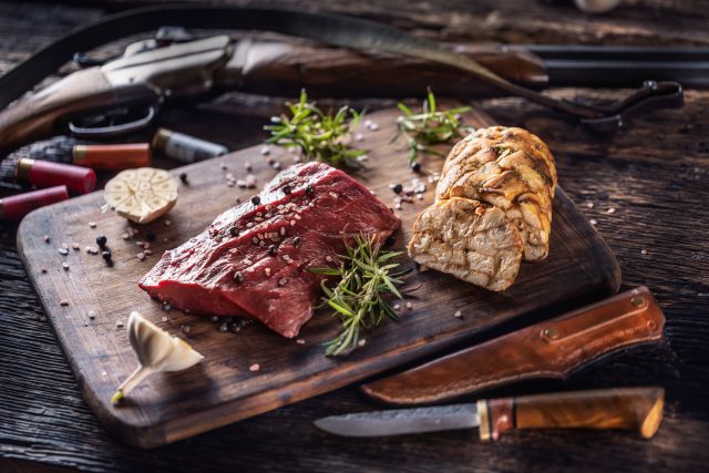 Nutrient rich raw deer venison prepared for a cooking process on a rustic wooden desk with roasted garlic, rosemary and huntig accesories like shot gun and ammunition.