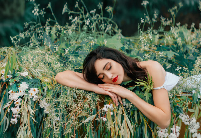 Young beauty sleeping woman resting in boat, decorated with willow branches, white flowers. Nymph girl closed eyes, enjoy silence summer nature river, water. Natural makeup red lips. Fantasy image