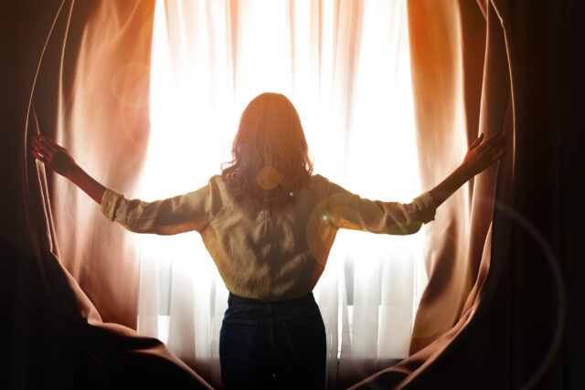 Woman opening window curtains at home, back view